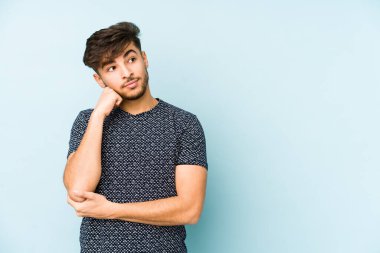 Hüzünlü ve dalgın hisseden, kopyalama alanına bakan, mavi bir arka planda izole edilmiş genç bir Arap..