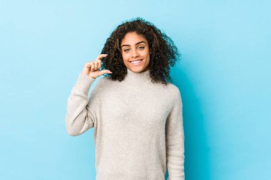 Genç Afro-Amerikan kıvırcık saçlı kadın işaret parmaklarıyla küçük bir şey tutuyor, gülümsüyor ve kendinden emin..