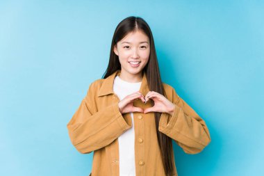 Mavi arka planda poz veren genç Çinli kadın izole edilmiş gülümseyen ve elleriyle kalp şeklini gösteren..