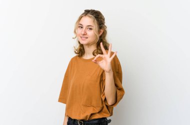 Beyaz arka plandaki genç beyaz kadın göz kırpıyor ve elleriyle iyi bir jest yapıyor..