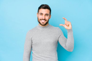 Mavi arka planda genç beyaz bir adam izole edilmiş, parmakları olan küçük bir şey tutuyor, gülümsüyor ve kendinden emin..