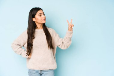 Mavi arka planda izole edilmiş genç Hintli kadın neşeli ve kaygısız parmakları olan bir barış sembolü gösteriyor..