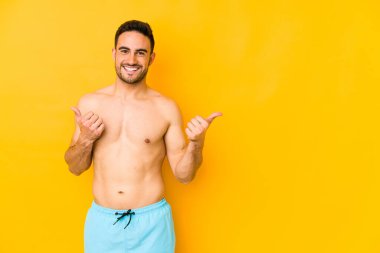 Sarı arka planda mayosu olan genç, beyaz bir adam. İki başparmağı da havada, gülümseyen ve kendine güveni tam beyaz bir genç..