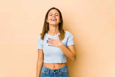 Bej arka planda izole edilmiş genç bir kadın yüksek sesle gülüyor elini göğsüne koyuyor..