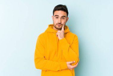 Mavi arka planda izole edilmiş genç beyaz adam şüpheli ve şüpheci bir ifadeyle yana bakıyor..