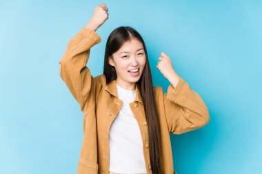 Mavi arka planda poz veren Çinli genç bir kadın özel bir günü kutluyor, atlıyor ve enerjiyle kollarını kaldırıyor..