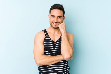 Mavi arka planda izole edilmiş genç beyaz adam gülümsüyor ve kendinden emin, elleriyle çenesine dokunuyor..