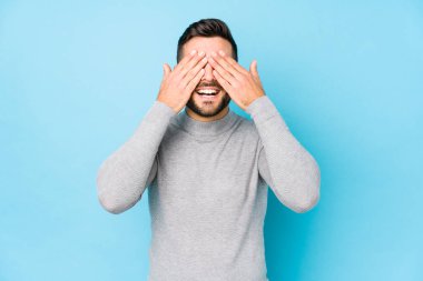 Mavi arka planda genç beyaz bir adam, izole edilmiş gözleri ellerle kaplıyor, geniş bir gülümsemeyle bir sürpriz bekliyor..