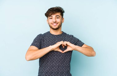 Mavi arka planda izole edilmiş genç bir Arap adam gülümsüyor ve elleriyle kalp şeklini gösteriyor..