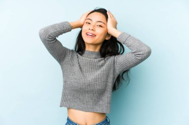 Mavi arka planda izole edilmiş genç Çinli kadın ellerini başının üstünde tutarak neşe içinde gülüyor. Mutluluk kavramı.