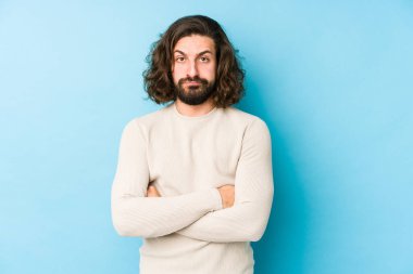 Uzun saçlı genç adam mavi bir arka planda izole edilmiş. Kameraya alaycı bir ifadeyle bakıyor..