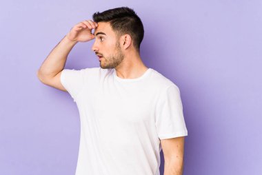 Mor arka planda izole edilmiş genç beyaz adam uzaklara bakıyor ve elini alnına koyuyor..