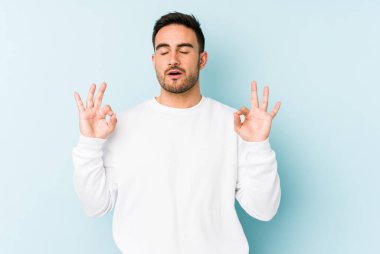 Mavi arka planda izole edilmiş genç beyaz adam yorucu bir günün ardından rahatlıyor. Yoga yapıyor..