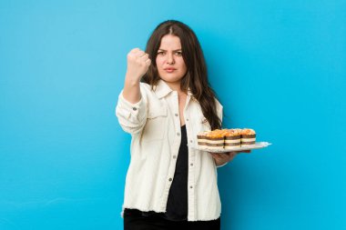 Kameraya yumruğunu gösteren küçük bir kek tutan kıvrımlı genç kadın, agresif yüz ifadesi..