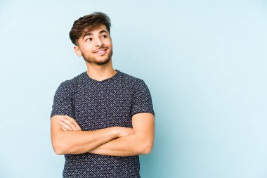Mavi bir arka planda izole edilmiş genç bir Arap adam kollarını kavuşturarak gülümsüyor..