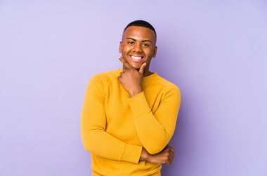 Mor arka planda izole olmuş genç Latin adam gülümsüyor ve kendinden emin, elleriyle çeneye dokunuyor..