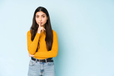 Genç Hintli kadın mavi arka planda izole edilmiş bir şekilde ya sır saklıyor ya da sessizlik istiyor..