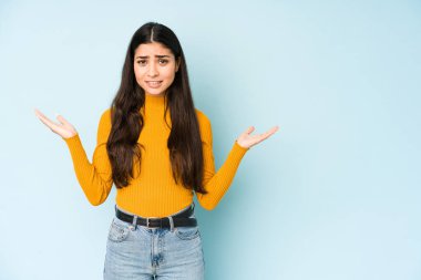 Mavi arka planda izole edilmiş genç bir Hintli kadın. Kafası karışmış ve omuz silkiyor. Kopya alanını tutmak için..