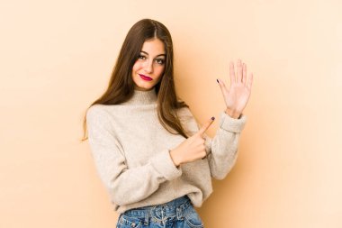 Beyazlı genç bir kadın bej arka planda izole edildi. Gülümseyerek beş numarayı parmaklarıyla gösterdi..