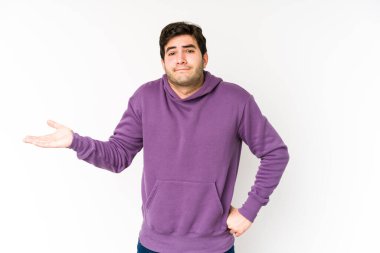 Beyaz arka planda izole edilmiş genç bir adam şüphe içinde ve sorgulama sırasında omuzlarını silkiyor..