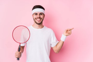 Badminton oynayan beyaz bir adam kendini soyutlamış gülümsüyor ve boş alanda bir şeyler gösteriyor..