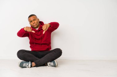 Yerde tek başına oturan genç latin adam baş parmağını aşağı gösteriyor ve hoşlanmadığını ifade ediyor..