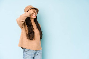 Mavi arka planda izole edilmiş bir şapka takan genç Hintli kadın elleriyle gözlerini kapatıyor, gülümseyerek bir sürpriz bekliyor..