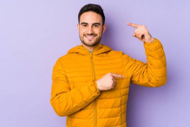 Mor arka planda izole edilmiş genç beyaz bir adam. Elinde işaret parmaklarıyla küçük bir şey tutuyor, gülümsüyor ve kendinden emin..