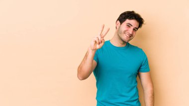 Bej arka planda izole edilmiş genç adam neşeli ve kaygısız parmakları olan bir barış sembolü gösteriyor..