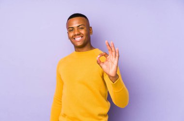 Mor arka planda izole edilmiş genç Latin adam neşeli ve kendinden emin, iyi bir jest yapıyor..