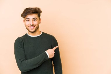 Bej bir arka planda izole edilmiş genç bir Arap adam gülümsüyor ve boş alanda bir şeyler gösteriyor..