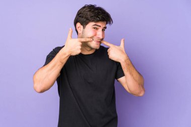 Mor arka planda izole olmuş genç adam gülümsüyor, parmaklarını ağıza doğrultuyor..