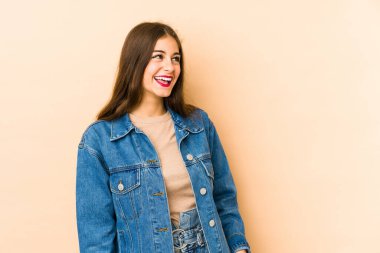 Beyaz tenli genç bir kadın bej arka planda izole edilmiş rahat ve mutlu bir şekilde gülüyor, boynunu uzatmış dişlerini gösteriyor..