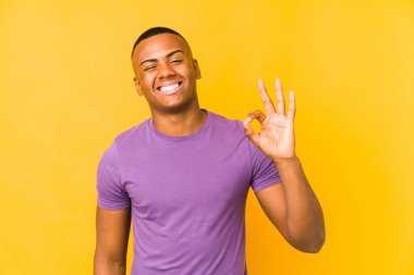 Sarı arka planda izole edilmiş genç Latin bir adam bir göz kırptı ve elleriyle iyi bir jest yaptı..