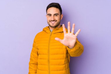 Mor arka planda izole edilmiş genç beyaz adam gülümseyerek beş numarayı parmaklarıyla gösteriyor..