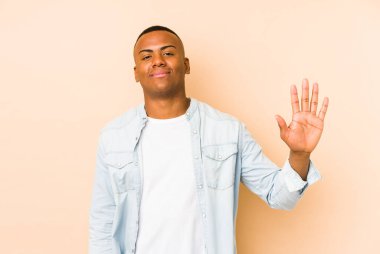 Genç latin adam bej arka planda izole edilmiş gülümsüyor beş numarayı parmaklarıyla gösteriyor..
