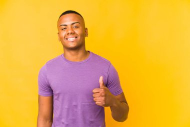 Sarı arka planda izole edilmiş genç latin adam gülümsüyor ve başparmağını kaldırıyor.