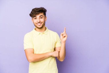 Mor arka planda izole edilmiş genç bir Arap adam işaret parmağıyla işaret ederek gülümsüyor..