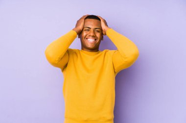 Mor arka planda izole olmuş genç latin adam mutlu bir şekilde ellerini başının üstünde tutuyor. Mutluluk kavramı.