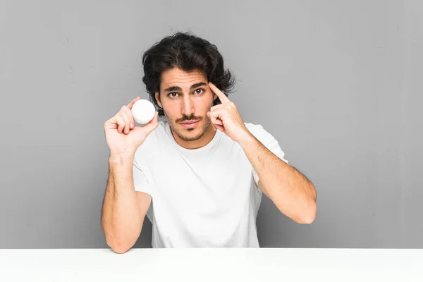 Joven Sosteniendo Una Crema Hidratante Apuntando Sien Con Dedo Pensando — Foto de Stock