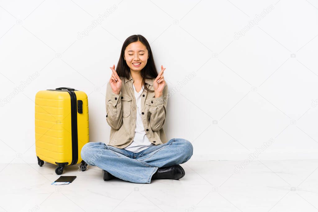 Young chinese traveler woman sitting holding a boarding passes crossing fingers for having luck