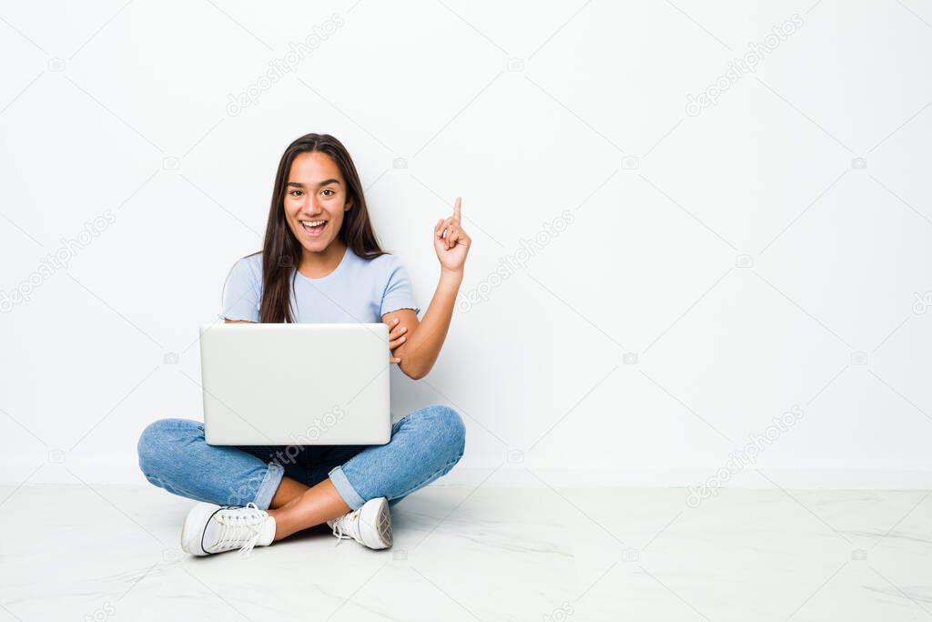 Young mixed race indian woman sitting working on laptop smiling cheerfully pointing with forefinger away.