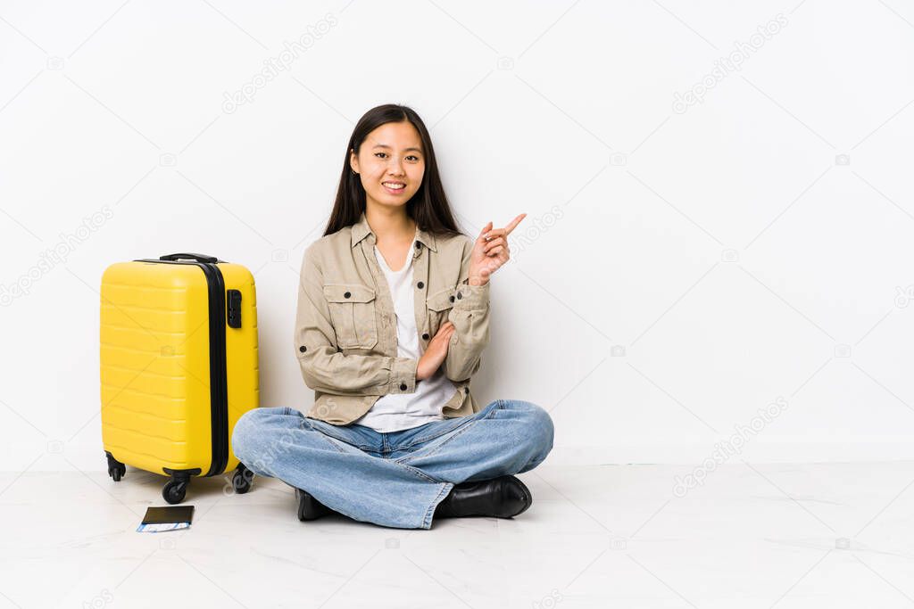 Young chinese traveler woman sitting holding a boarding passes smiling cheerfully pointing with forefinger away.