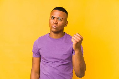 Sarı arka planda izole edilmiş genç latin adam kameraya yumruk gösteriyor, agresif yüz ifadesi..