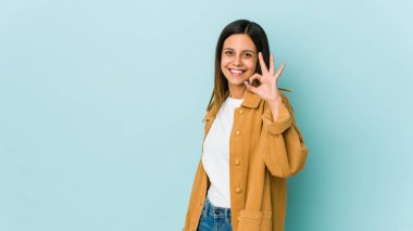 Mavi arka planda izole edilmiş genç bir kadın bir göz kırpıyor ve elleriyle iyi bir jest yapıyor..