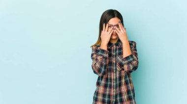 Mavi arka planda izole edilmiş genç bir kadın kameraya parmaklarıyla göz kırpıyor, yüzünü kapatmaktan utanıyor..