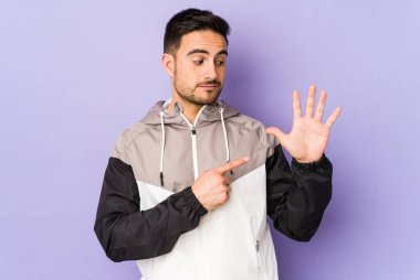 Mor arka planda izole edilmiş genç beyaz adam gülümseyerek beş numarayı parmaklarıyla gösteriyor..