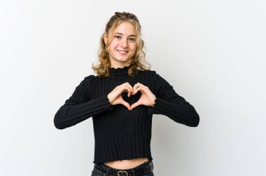 Beyaz arka plandaki genç beyaz kadın gülümsüyor ve elleriyle kalp şeklini gösteriyor..