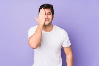 Mor arka planda izole edilmiş genç bir adam yüzünün yarısını avuç içiyle kaplayarak eğleniyor..
