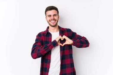 Beyaz arka planda poz veren beyaz tenli genç bir adam izole edilmiş, gülümseyen ve elleriyle kalp şeklini gösteren..